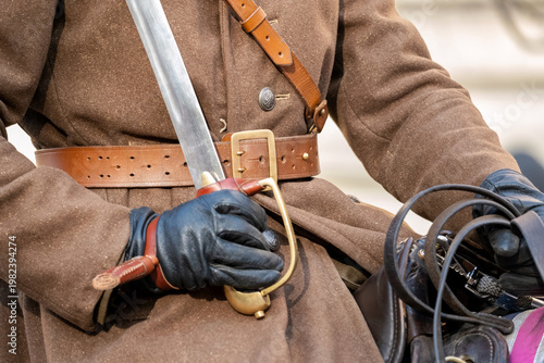 Close-up of a historical cavalry reenactor in brown traditional military uniform holding a curved saber while seated on horseback, gloved hands, leather belt reins detail during outdoor heritage event