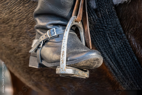 Extreme close-up of a cavalry rider’s black leather boot in a metal stirrup on horseback, traditional mounted uniform detail with horse tack and dark mane at a historical reenactment