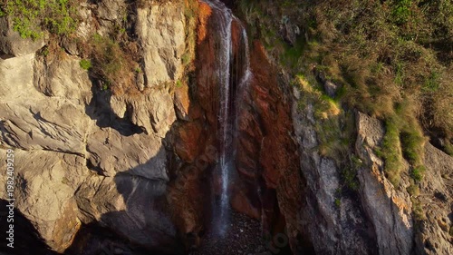 Aerial view of waterfall cascading down rocky cliff directly into ocean. Dramatic coastal landscape with waves, nature scenery and exotic tropical style environment. High quality 4k footage