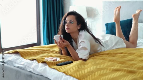 Hot Caucasian girl in glasses and lying on her bed, watching tv with interest, drinking coffee from white cup and eating biscuit. Beautiful woman eating chocolate, and moving her legs in her bedroom.