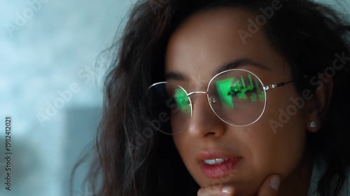 Caucasian female with long brown hair is wearing glasses, lying on bed and watching movie on her computer. Pretty young woman relaxing and looking at screen with interest. Real-time video, close up.