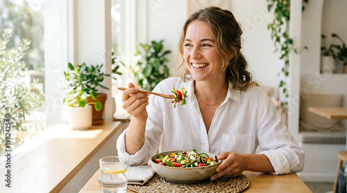 Woman eating healthy salad with chopsticks by the window in a bright room