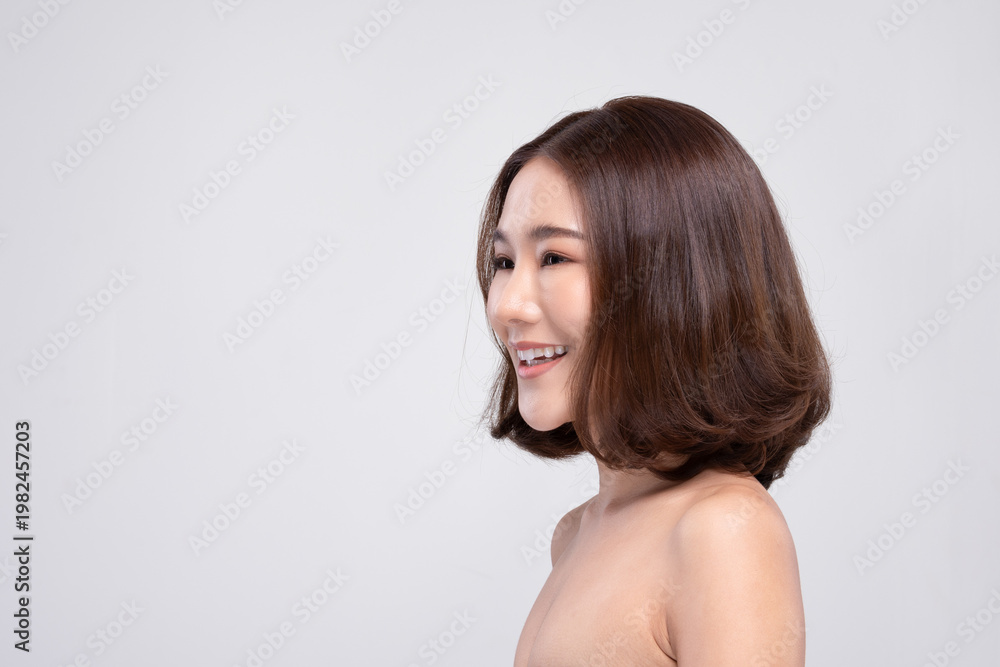 Obraz premium Beautiful young asian woman smiling happily with bare shoulder showing perfect skin and short brown hair enjoying facial skincare beauty treatment isolated on white studio background
