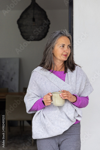 Senior woman standing in doorway in gray wrap holding ceramic mug looking toward dining table