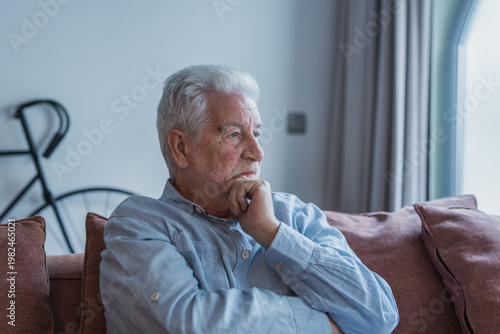 An elderly man sits on a sofa with his hand on his chin, gazing out a window in a softly lit living room and conveying quiet reflection, nostalgia and calm solitude at home.