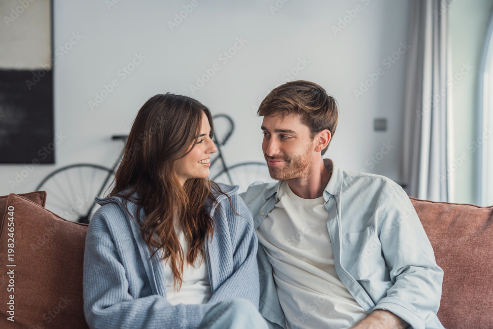 Obraz premium A young couple relaxes on a sofa, smiling and sharing a warm, intimate moment at home. Casual clothing, soft lighting and a bicycle in the background create a comfortable lifestyle scene