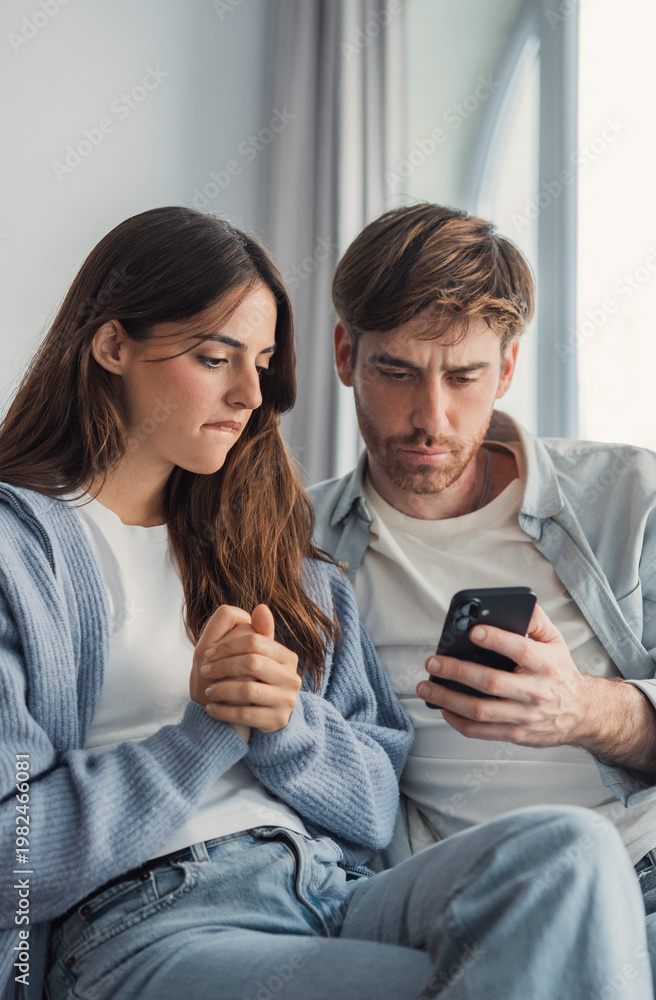 Fototapeta premium A young couple sits close on a sofa, intently reading a phone with worried expressions; conveys concern, tension, communication issues and emotional uncertainty in a domestic setting