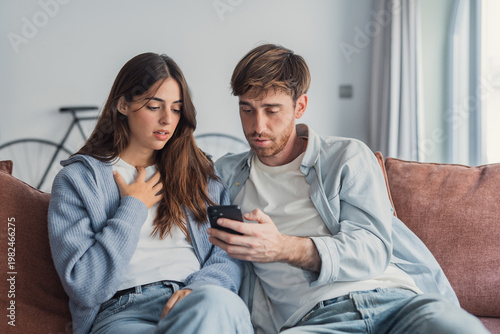 Foto A young couple sits close on a living room sofa, reacting with surprise as they