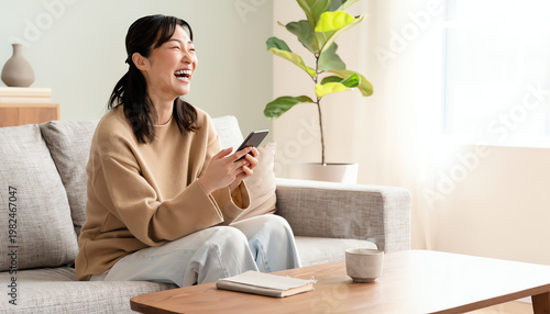 A woman sitting on a sofa, laughing joyfully while holding a smartphone in a bright, cozy living room.
