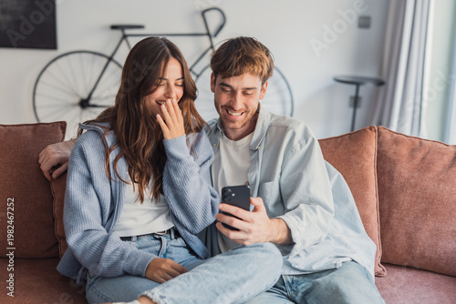 Photography A young couple relaxes on a cozy sofa at home, laughing and sharing a moment while looking at a smartphone