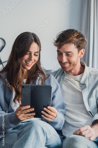 A happy young couple sits close on a couch, sharing a tablet and enjoying digital content together. Warm, relaxed mood conveys intimacy, modern technology use, and comfortable home life