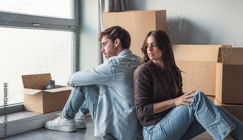 Quadro em tela A young couple sits back-to-back among cardboard moving boxes by a window, appearing distant and tense after a move