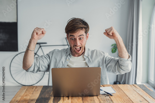 Quadro su tela Young man raises his fists in excitement while looking at a laptop on a wooden table, celebrating success during remote work