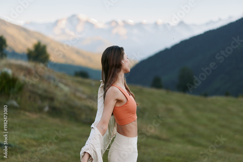 A relaxed woman stands outdoors in a mountain landscape, gazing upward with a light breeze in her hair, wearing an orange top and white skirt, exuding freedom and quiet wonder.