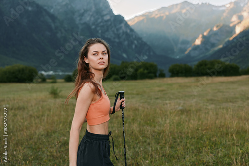 A young woman stands in a meadow with distant mountains, wearing an orange crop top and black shorts, holding a trekking pole, gazing into the distance with a calm, confident expression.