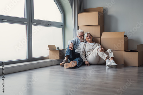 An older couple sits on the floor smiling among packed cardboard boxes, enjoying a calm moment while unpacking and celebrating their move into a bright new home together.