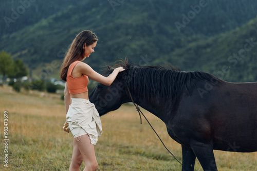 A relaxed woman in casual attire stands beside a calm horse in a wide meadow, gently reaching to pet its neck. The tranquil countryside setting conveys connection, care, and a peaceful outdoor