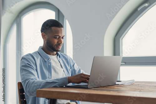 A young man types on a laptop at a wooden desk in a bright home office, focused and calm while working remotely. Natural light, casual attire and a clean minimalist workspace convey productivity 