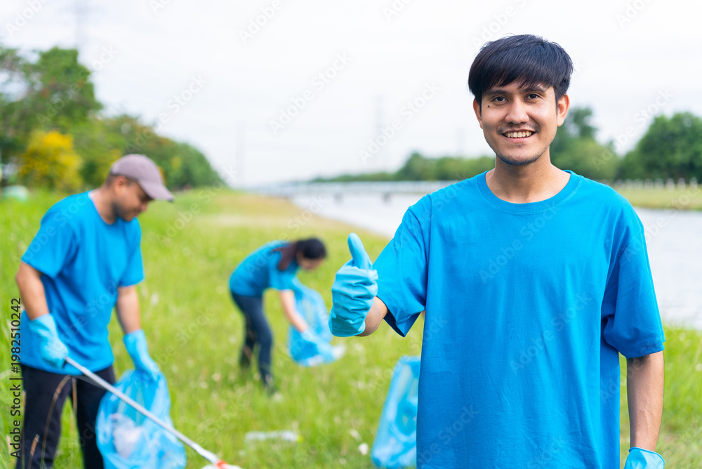 Naklejka premium Volunteering, charity and clean environment concept. Male volunteers wearing blue t-shirt and protection glove standing and thumbs up to camera with volunteers diverse group cleaning park background.