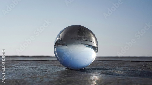 A crystal globe sits on a rocky beach by the sea in the morning. The blue sky and reflect the sunlight, creating an image resembling a world map within the globe, the  interconnectedness of the globe.