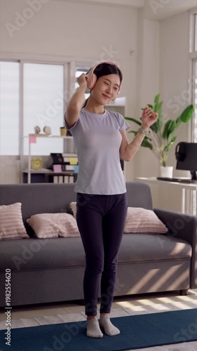 Young Asian woman dancing wearing headphones enjoying music indoor home living room for relaxation happiness and stress relief wellbeing fitness activity