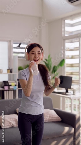 Asian young adult woman enjoying music singing into water bottle headphones home modern living room leisure lifestyle relaxation entertainment self care activity