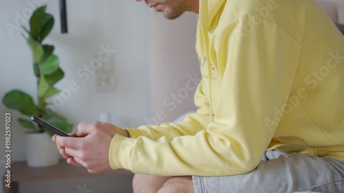 Adult man in yellow hoodie using smartphone while sitting on bed in home interior. Concept of mobile communication online activity and everyday technology use