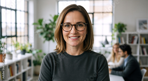 Smiling businesswoman with glasses looking at camera in modern office. Confident female manager portrait. Corporate team working in blurred background