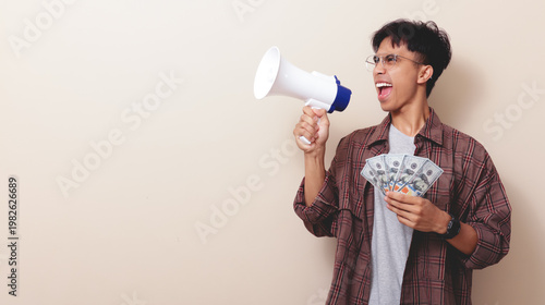 Fotografía Excited young Asian man holding money banknotes and shouting into a megaphone on isolated background