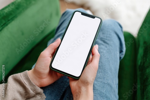 A person comfortably sits on a green couch, holding a smartphone with a blank screen, suggesting a moment of relaxation and digital engagement.