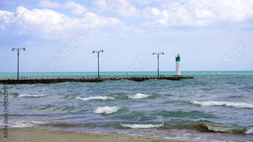 
A small white lighthouse with a green top stands at the end of a stone pier extending into the rolling turquoise waves of Lake Ontario along the Oshawa lakefront