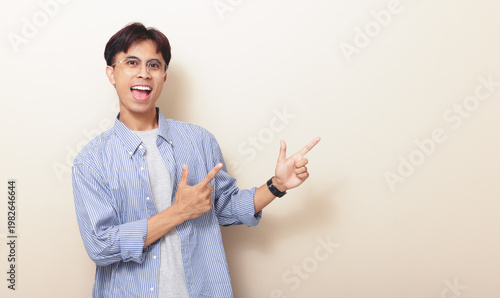 Obraz na plátně Happy excited young Asian man pointing finger to copy space area in sideways on isolated background