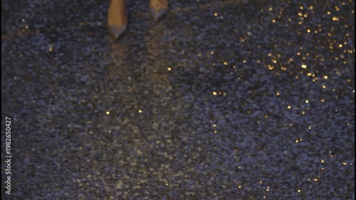 A woman's legs wearing metallic high heels and sequined pants on a glowing asphalt road at night.
