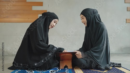 Two Muslim women in black hijab reading Quran together while sitting on a prayer mat in a calm indoor environment, representing faith, learning, and spiritual connection.
