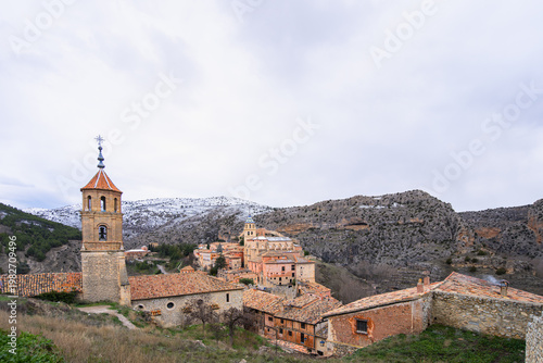 A scenic view of the historic medieval town of Albarracin in Spain, featuring traditional stone buildings and a church tower against snowy mountains.