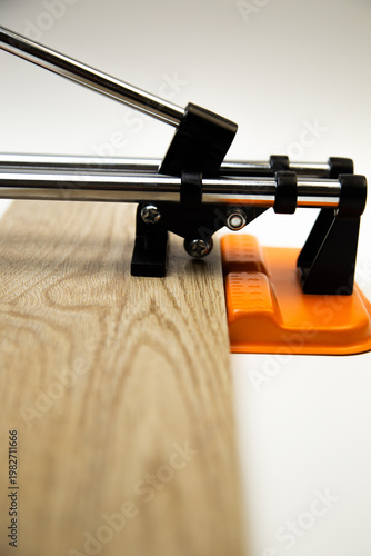 Close-up side view of an orange mechanical tile cutter, which fixed ceramic tiles of wooden colors, above it is a cutting disc on a white background. The concept of renovation, tiling, construction