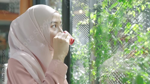 Muslim woman in hijab enjoying coffee near a window with greenery, representing relaxation, lifestyle, and modern daily life.
