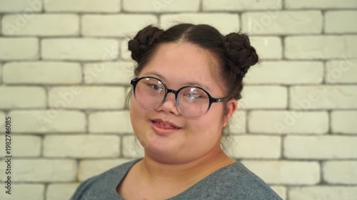 Close-up portrait of a young woman with Down syndrome wearing glasses and smiling, isolated against light brick wall background
