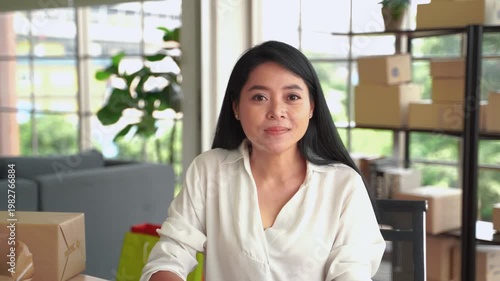 Confident woman business owner smiling at camera while working in home office with boxes and supplies for online store
