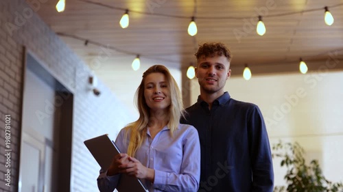 Happy two young colleagues standing in cafe. Lady holding laptop. Couple posing at camera. Lifestyle, partnership, job concept. Real time
