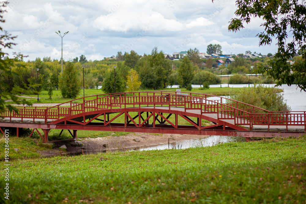 Fototapeta premium Red wooden bridge over a small river surrounded by lush greenery and trees under a cloudy sky in a tranquil park setting