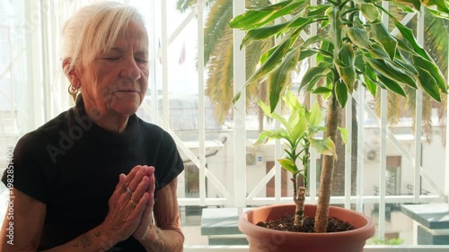 Serious senior woman praying or meditating with joined hands on a balcony. Authentic portrait of elderly spirituality and mindfulness