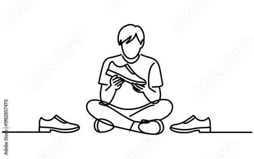 A man sits cross-legged on the floor, reading a book with shoes nearby him.