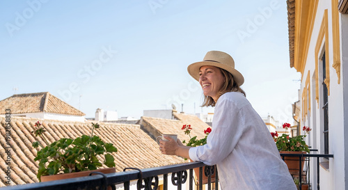 A woman wearing a straw hat and a white shirt smiles as she holds a mug on a balcony overlooking rooftops.