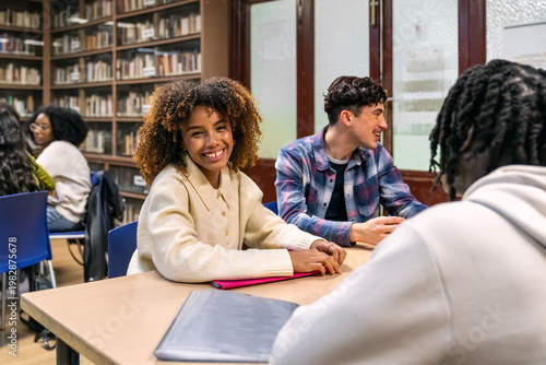 Young female student smiling while studying with classmates and teacher at a table in a university library
