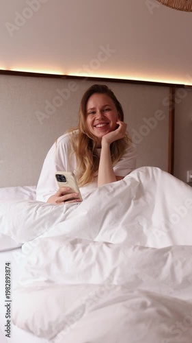 Happy young woman lying in white bed using smartphone, smiling at camera and blowing a kiss. Perfect for lifestyle, morning routine, social media and positive emotion themed projects