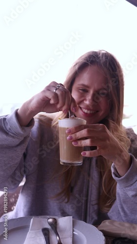 Happy young woman stirring latte coffee, tasting and licking spoon with joyful smile at cafe. Perfect for lifestyle, coffee, cafe culture, positive emotion and beverage themed projects
