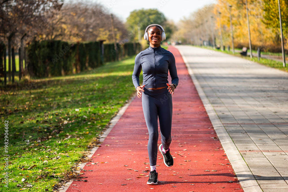 Obraz premium Happy black woman is jogging in the city park on sunny day. Woman in athletic wear is having outdoor workout.