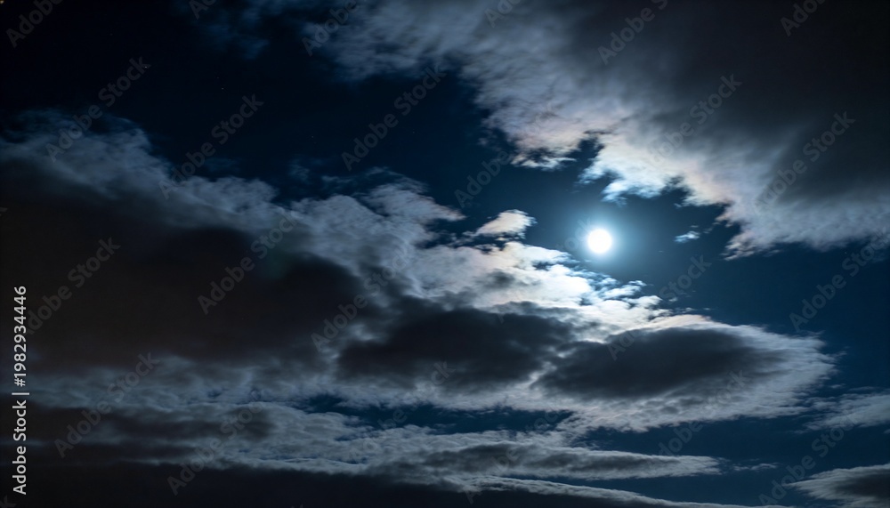 Obraz premium Glowing nearly full moon backlighting layered clouds at night, showing silver linings and halo