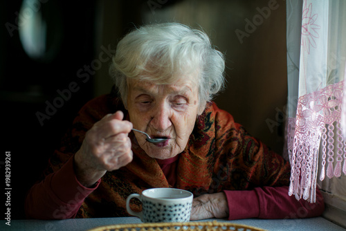 Elderly woman eating from a cup with a spoon near a window, soft natural light, quiet domestic interior scene.